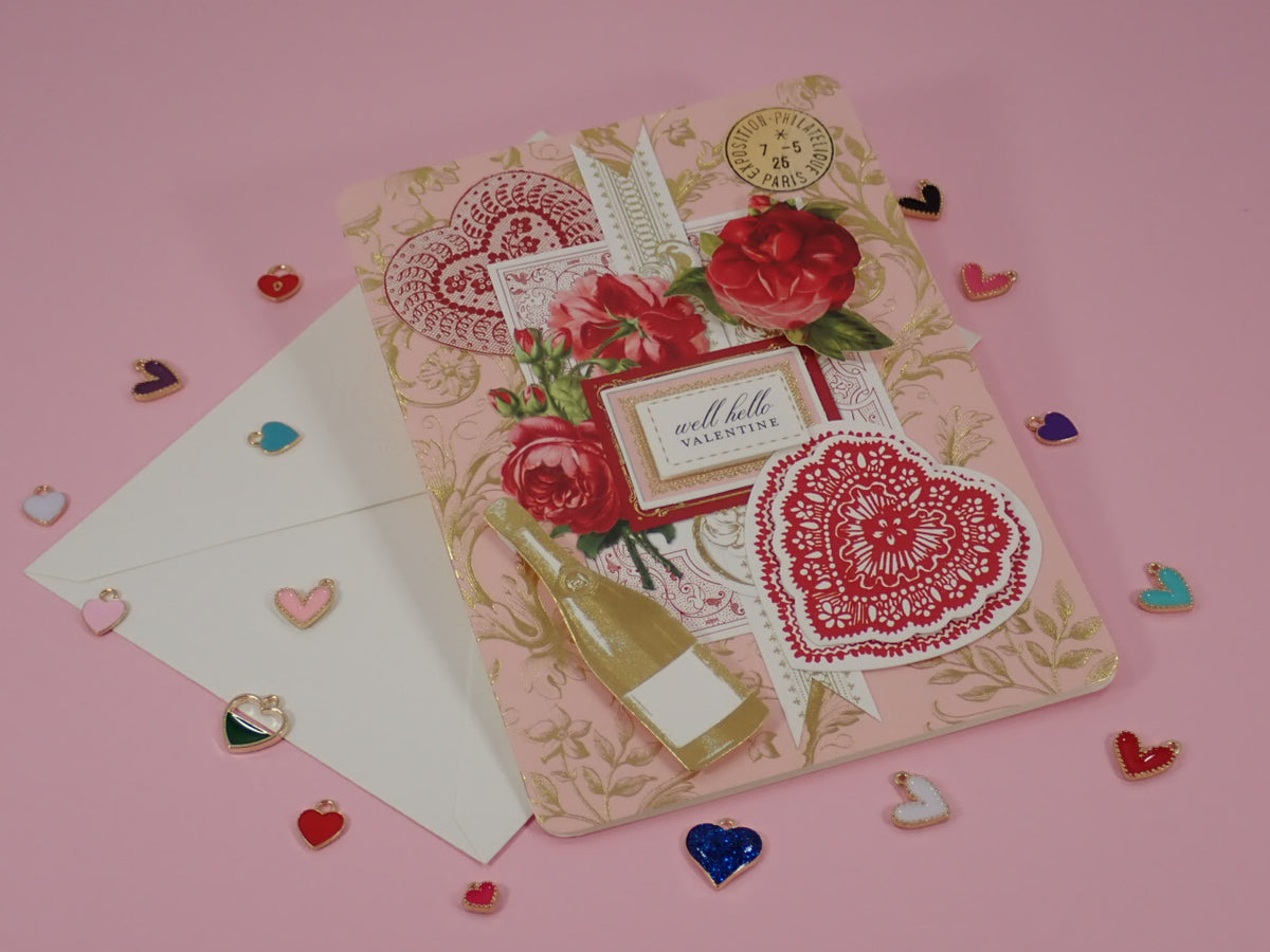 A decorative handcrafted Valentine's Day, greeting card with a vintage Victorian  theme, featuring red roses, lace hearts, a champagne bottle illustration, and a small label reading 'well hello valentine', displayed on a pink background with a white envelope and scattered heart-shaped charms.