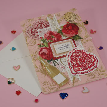 A decorative handcrafted Valentine's Day, greeting card with a vintage Victorian  theme, featuring red roses, lace hearts, a champagne bottle illustration, and a small label reading 'well hello valentine', displayed on a pink background with a white envelope and scattered heart-shaped charms.