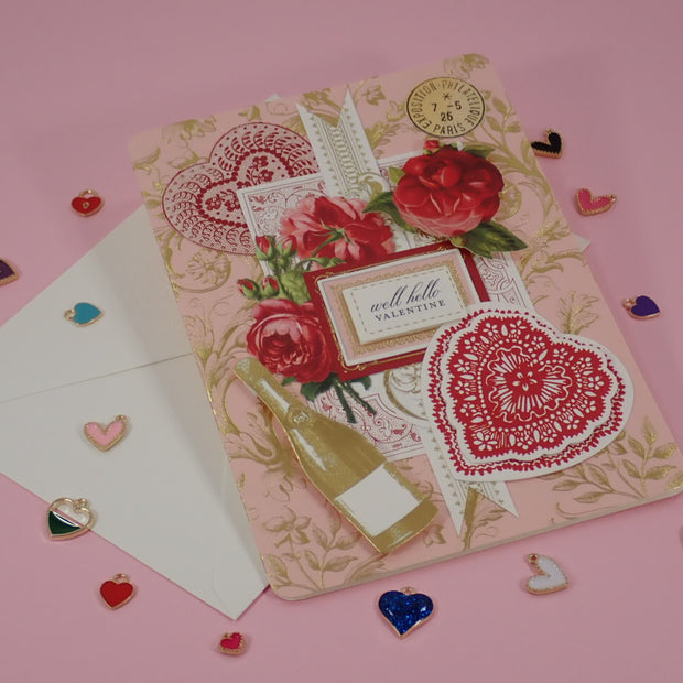 A decorative handcrafted Valentine's Day, greeting card with a vintage Victorian  theme, featuring red roses, lace hearts, a champagne bottle illustration, and a small label reading 'well hello valentine', displayed on a pink background with a white envelope and scattered heart-shaped charms.