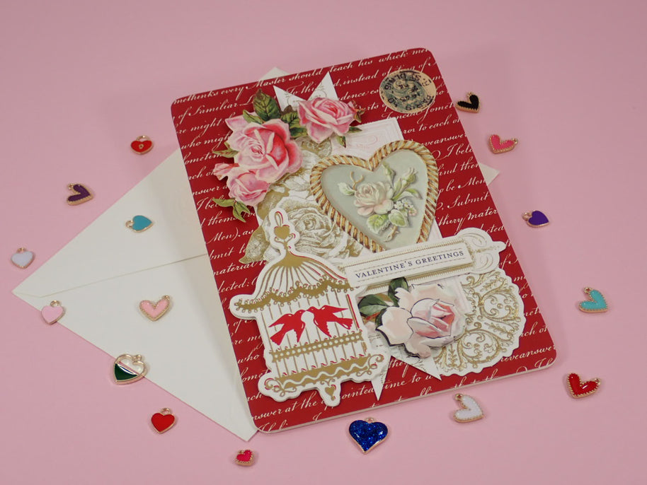 A handcrafted, Valentine's Day greeting card with a red patterned background, layered with pink roses, a birdcage motif, a heart-shaped floral emblem with a white envelope.