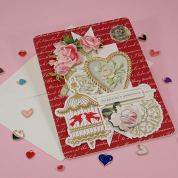 A handcrafted, Valentine's Day greeting card with a red patterned background, layered with pink roses, a birdcage motif, a heart-shaped floral emblem with a white envelope.