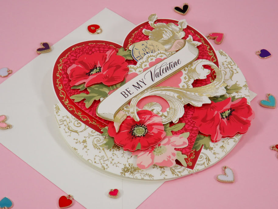 A 3D layered, handcrafted Valentine's Day card with red poppy flowers, gold filigree, and a white banner reading 'BE MY Valentine', displayed on a pink background with scattered heart-shaped confetti.