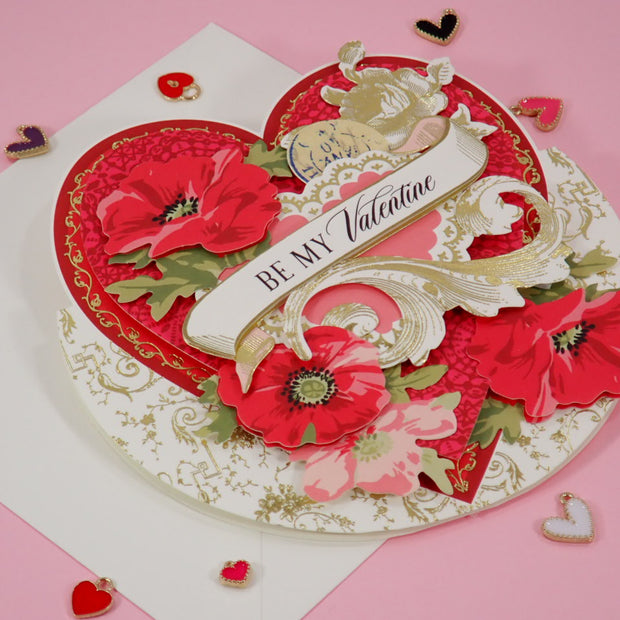 A 3D layered, handcrafted Valentine's Day card with red poppy flowers, gold filigree, and a white banner reading 'BE MY Valentine', displayed on a pink background with scattered heart-shaped confetti.
