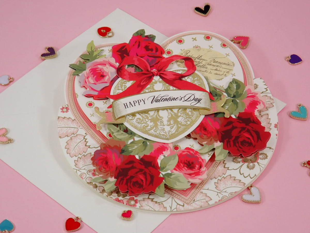 A handcrafted, 3D layered Valentine's Day card with a heart design, featuring red roses, green leaves, a red ribbon bow, and a central banner reading 'Happy Valentine's Day' on a pink background.