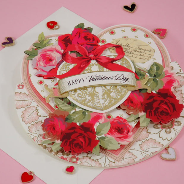 A handcrafted, 3D layered Valentine's Day card with a heart design, featuring red roses, green leaves, a red ribbon bow, and a central banner reading 'Happy Valentine's Day' on a pink background.