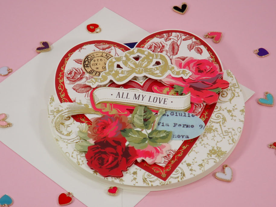 A handcrafted heart-shaped layered Valentine's Day card with red roses, gold filigree, a white ribbon reading 'ALL MY LOVE', and a small blue handwritten note, displayed on a pink background with scattered heart-shaped confetti.