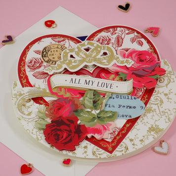 A handcrafted heart-shaped layered Valentine's Day card with red roses, gold filigree, a white ribbon reading 'ALL MY LOVE', and a small blue handwritten note, displayed on a pink background with scattered heart-shaped confetti.