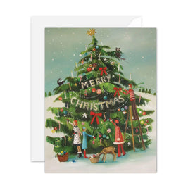 An artisan made, Christmas greeting card featuring an illustrated Christmas tree decorated with ornaments, lights, and a banner that reads 'Merry Christmas.' Two children, a dog, and a ladder are part of the artwork, with a white envelope behind the card on a light gray background.