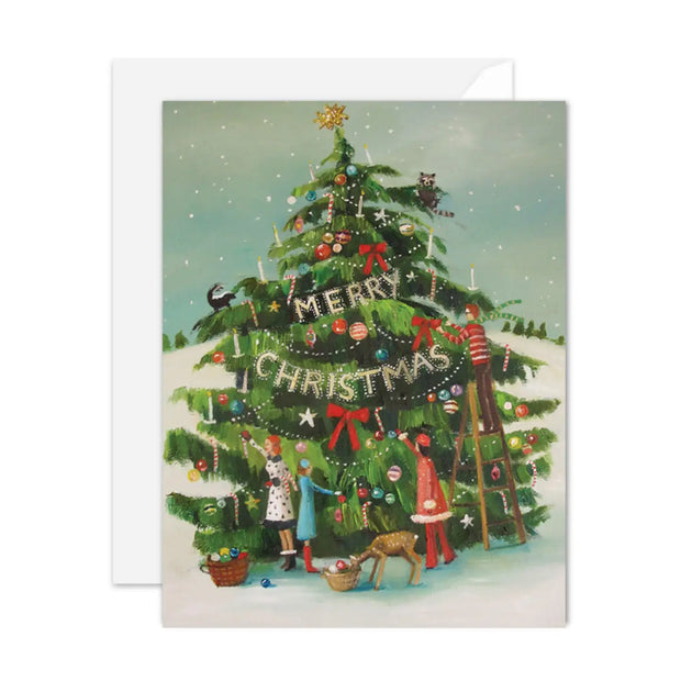 An artisan made, Christmas greeting card featuring an illustrated Christmas tree decorated with ornaments, lights, and a banner that reads 'Merry Christmas.' Two children, a dog, and a ladder are part of the artwork, with a white envelope behind the card on a light gray background.