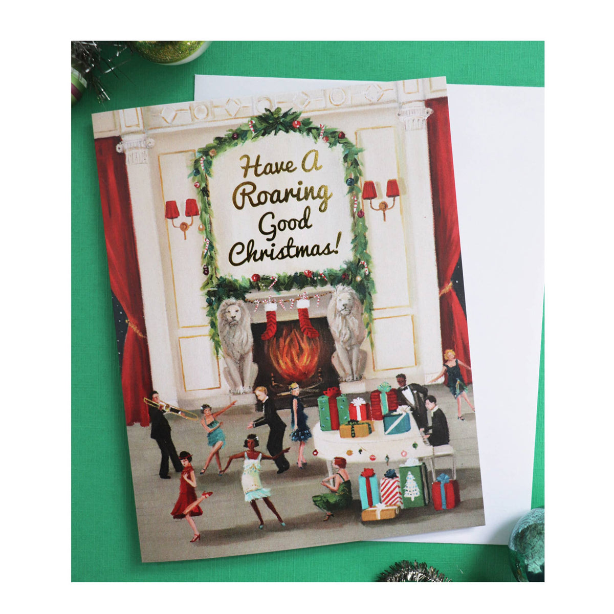 An artisan made, Christmas greeting card featuring a festive fireplace scene with the text 'Have A Roaring Good Christmas!' in gold script. The card is displayed on a green surface next to a white envelope.