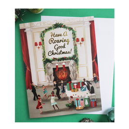 An artisan made, Christmas greeting card featuring a festive fireplace scene with the text 'Have A Roaring Good Christmas!' in gold script. The card is displayed on a green surface next to a white envelope.