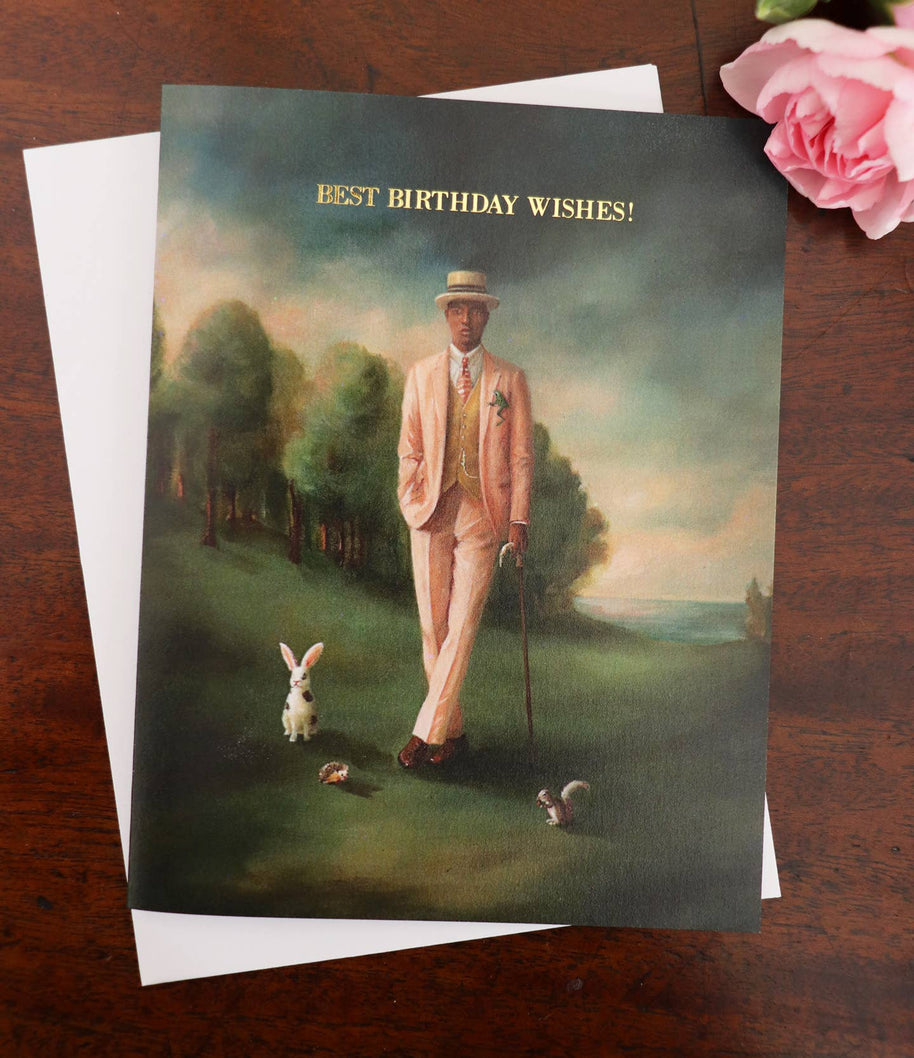 An artisan made birthday greeting card featuring a whimsical painting of a man in a light pink suit walking with a cane in a landscape, with a white envelope behind it on a dark wooden surface and a pink rose in the corner. The card reads 'BEST BIRTHDAY WISHES!'.