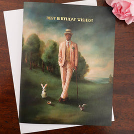 An artisan made birthday greeting card featuring a whimsical painting of a man in a light pink suit walking with a cane in a landscape, with a white envelope behind it on a dark wooden surface and a pink rose in the corner. The card reads 'BEST BIRTHDAY WISHES!'.