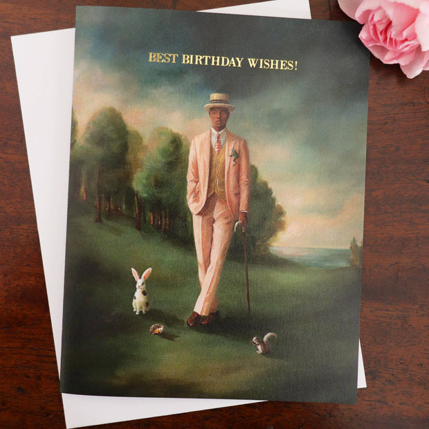An artisan made birthday greeting card featuring a whimsical painting of a man in a light pink suit walking with a cane in a landscape, with a white envelope behind it on a dark wooden surface and a pink rose in the corner. The card reads 'BEST BIRTHDAY WISHES!'.