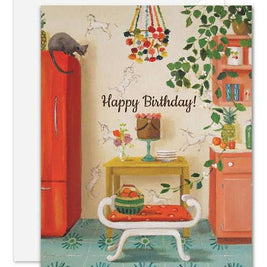 A vintage-style illustrated artisan made, birthday greeting card with a white envelope behind it. The card shows a retro kitchen scene with a red refrigerator, green leafy vines, a small bench with an orange cushion, a cake stand, and the words 'Happy Birthday!' in script.