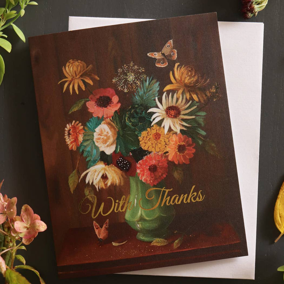 An artisan made Thank You greeting card with a vintage-style floral bouquet in a green vase and the words 'With Thanks' in gold script, placed on top of a white envelope against a dark tabletop with decorative flowers around the edges.